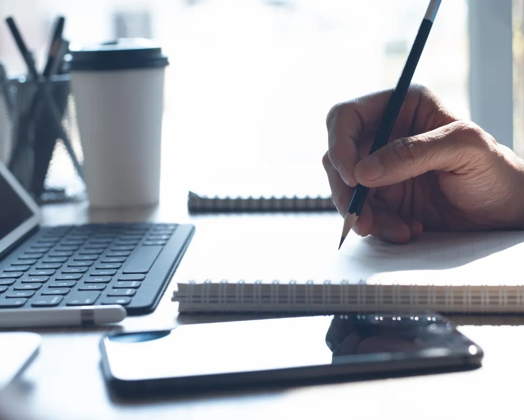 Hand schreibt mit Bleistift in einen Spiralblock, daneben Tastatur, Smartphone und Kaffeebecher